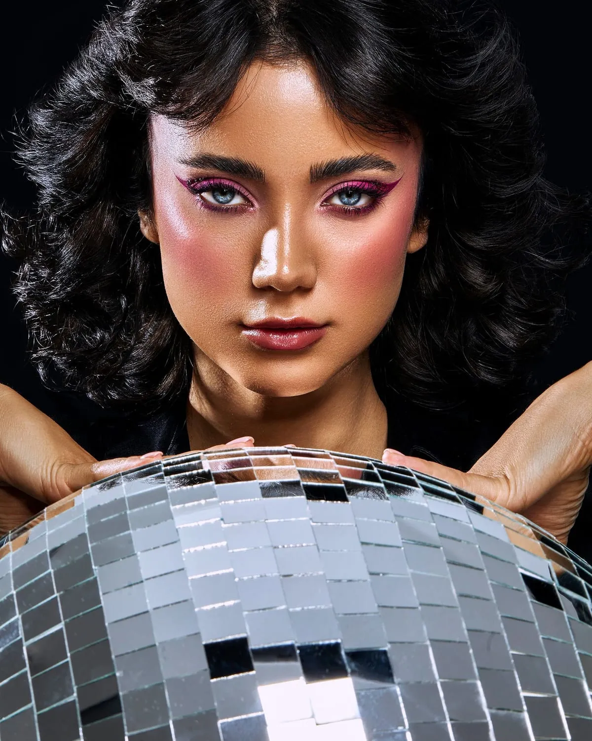 Woman with 70s disco makeup and voluminous curls leaning on a mirror ball